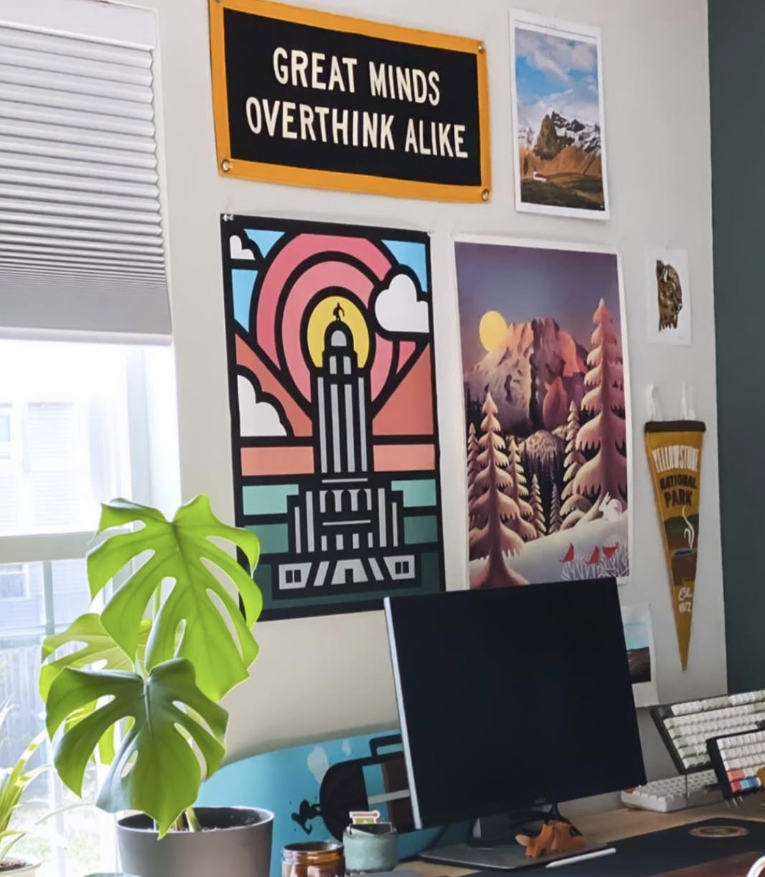 Studio wall with a 'Great minds overthink alike' pennant, screen prints and a monstera plant